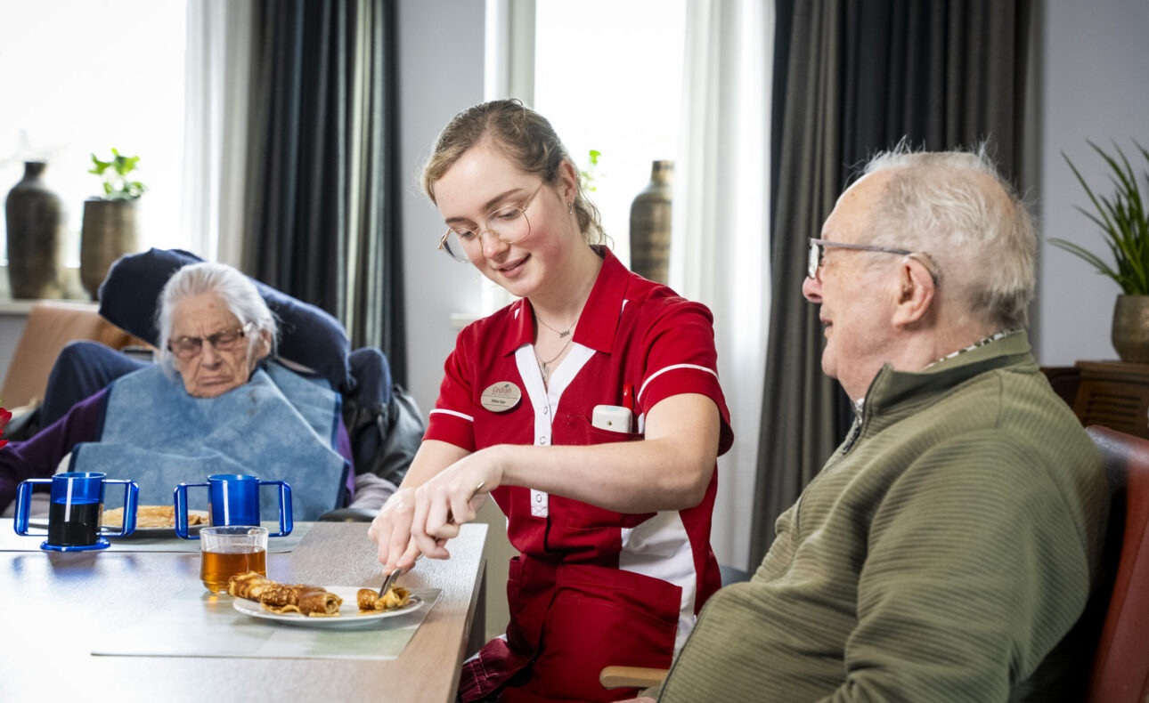 Een zorgmedewerker in rood uniform helpt een oudere man aan tafel. Een andere oudere persoon zit naast hen in een rolstoel.