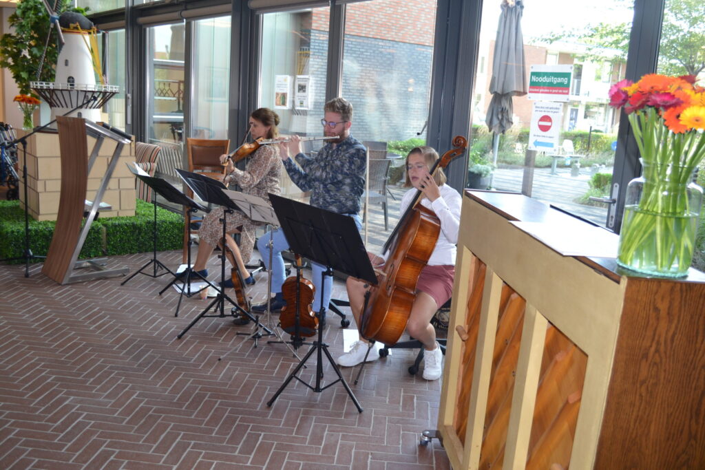 Drie muzikanten spelen viool, cello en harp in een lichte ruimte met grote ramen. Op de voorgrond staat een piano.