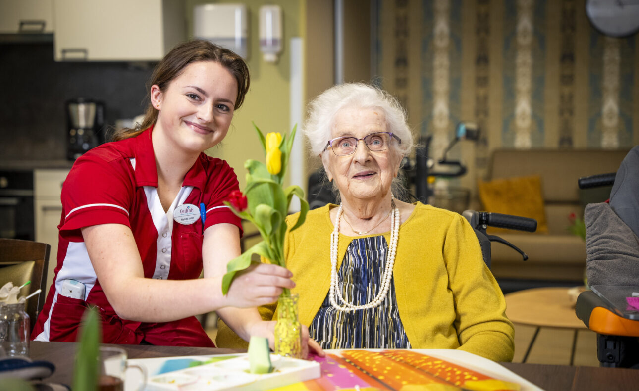 Een jonge zorgmedewerker in rood uniform geeft een gele tulp aan een oudere vrouw met een bril en gele trui in een woonkamer.