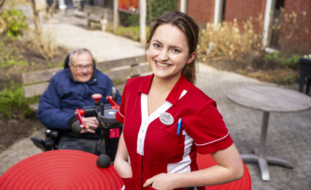 Een zorgmedewerker in een rood uniform met een cliënt op de achtergrond.