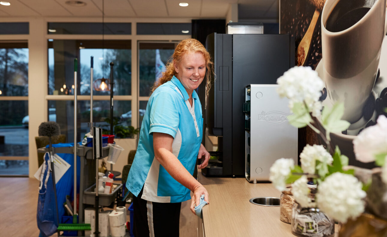 Een huishoudelijk medewerker in een blauw uniform. Schoonmaakkar achter zich en maakt schoon.